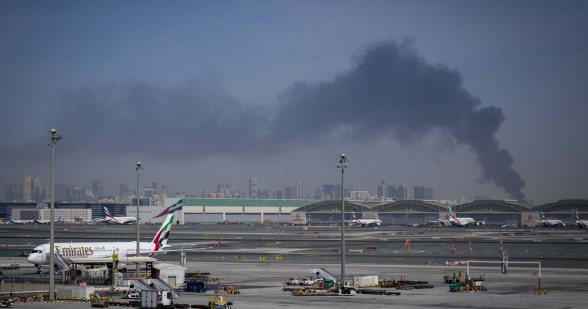 A plume of smoke caused by an Iranian strike is seen in the background as Emirates planes are parked at Dubai International Airport after its closure in Dubai, United Arab Emirates, Sunday, March 1, 2026 (AP Photo/Altaf Qadri)